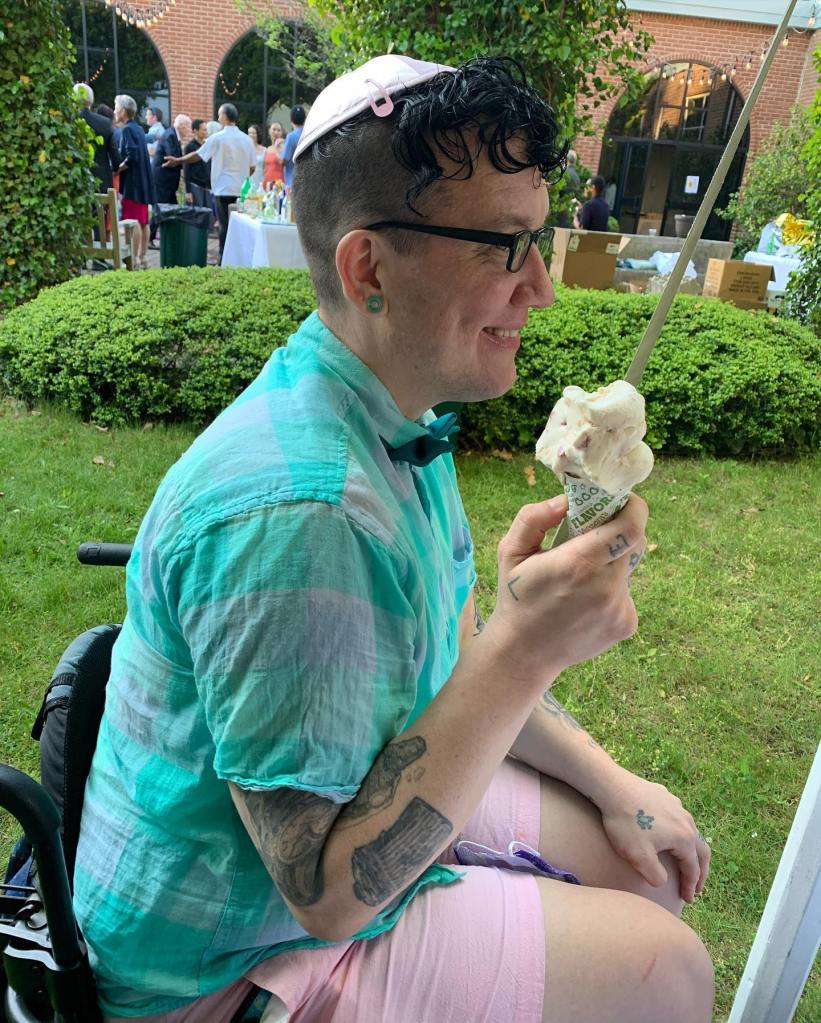 A side view of Mo Netz sitting in his wheelchair wearing a kippah and holding an ice cream cone.